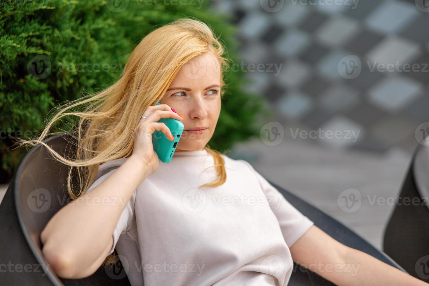 Young blond woman sittings outdoors in the city and making a call with her smartphone. Girl talking with friends with mobile phone. Leisure activity, communication. Conversation. Mobile technology. photo