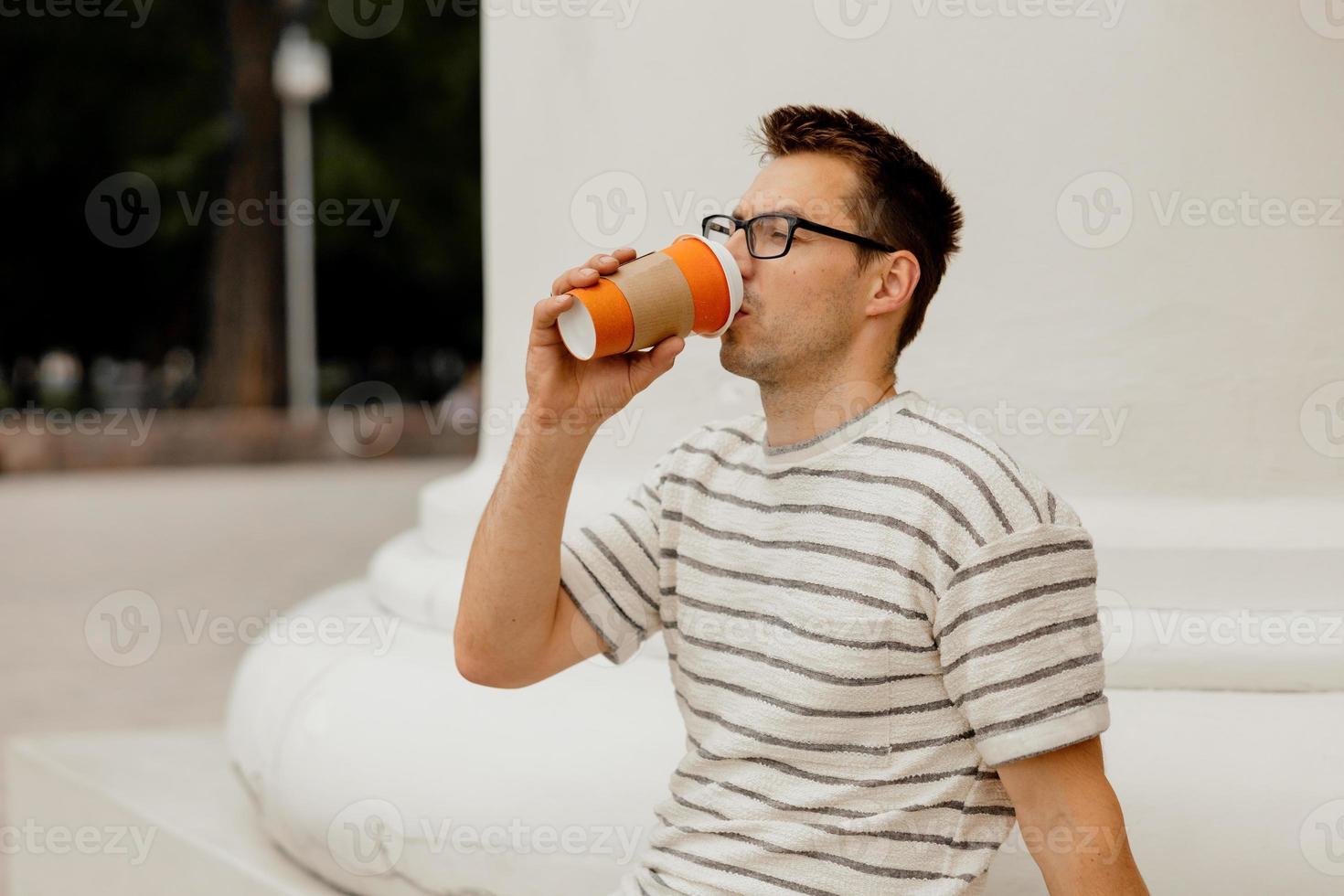 hombre adulto joven sentado al aire libre, tomando café, disfrutando del buen tiempo y la vista de la ciudad. el hombre tiene un descanso del trabajo, pasa tiempo al aire libre y se relaja. tiempo contigo mismo, soñar, salud mental. foto