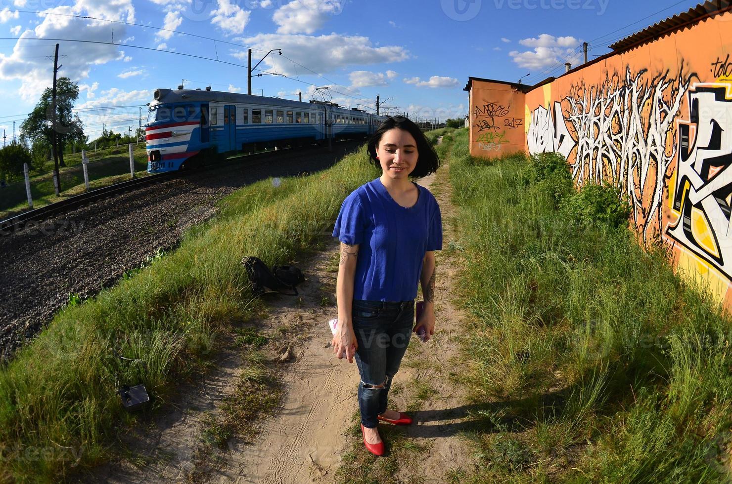 Portrait of an emotional young girl with black hair and piercings. A wide-angle photo of a girl with aerosol paint cans in the hands on a graffiti wall background. A modern portrait of a fisheye lens