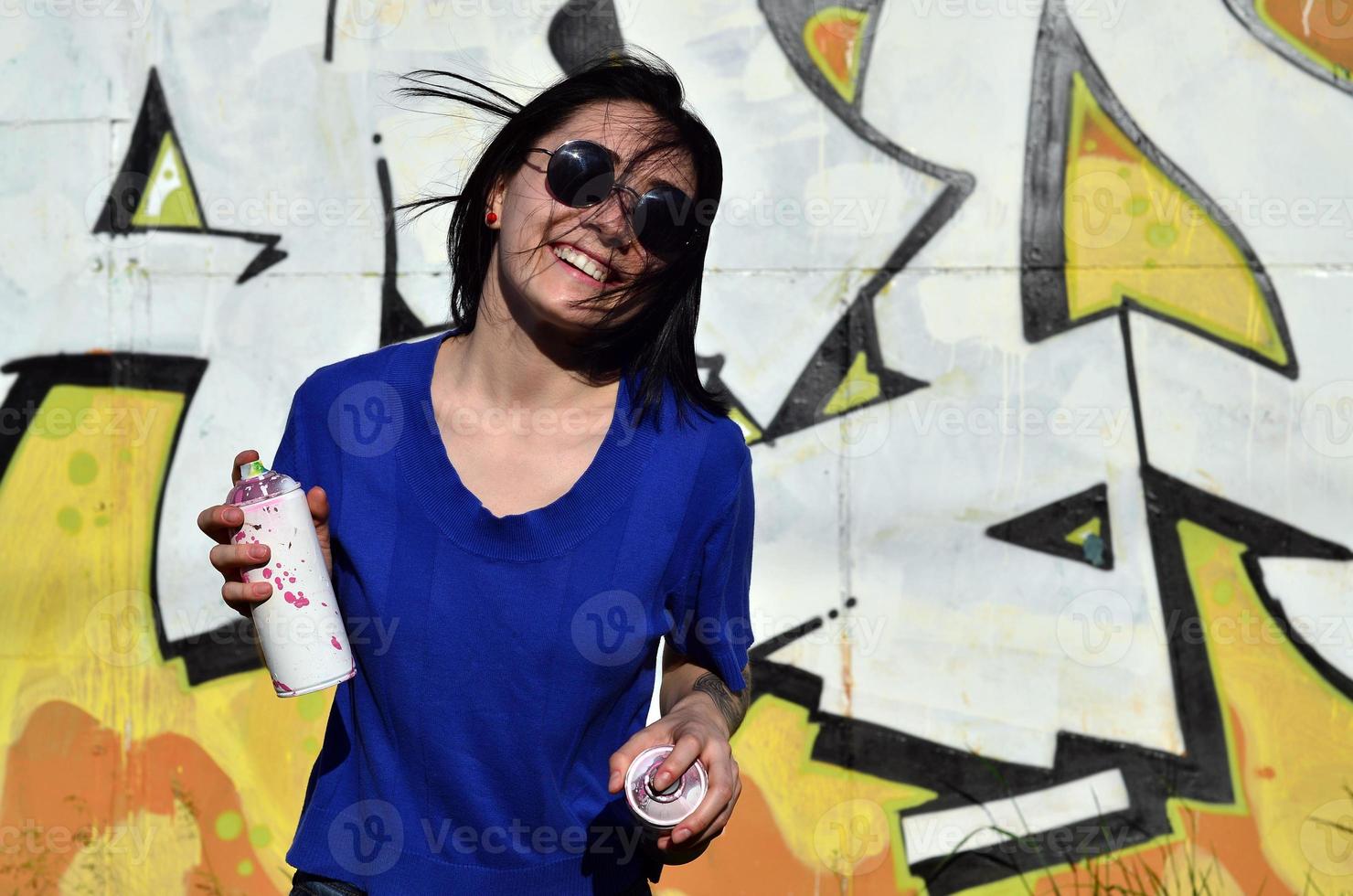 Portrait of an emotional young girl with black hair and piercings. Photo of a girl with aerosol paint cans in hands on a graffiti wall background. The concept of street art and use of aerosol paints