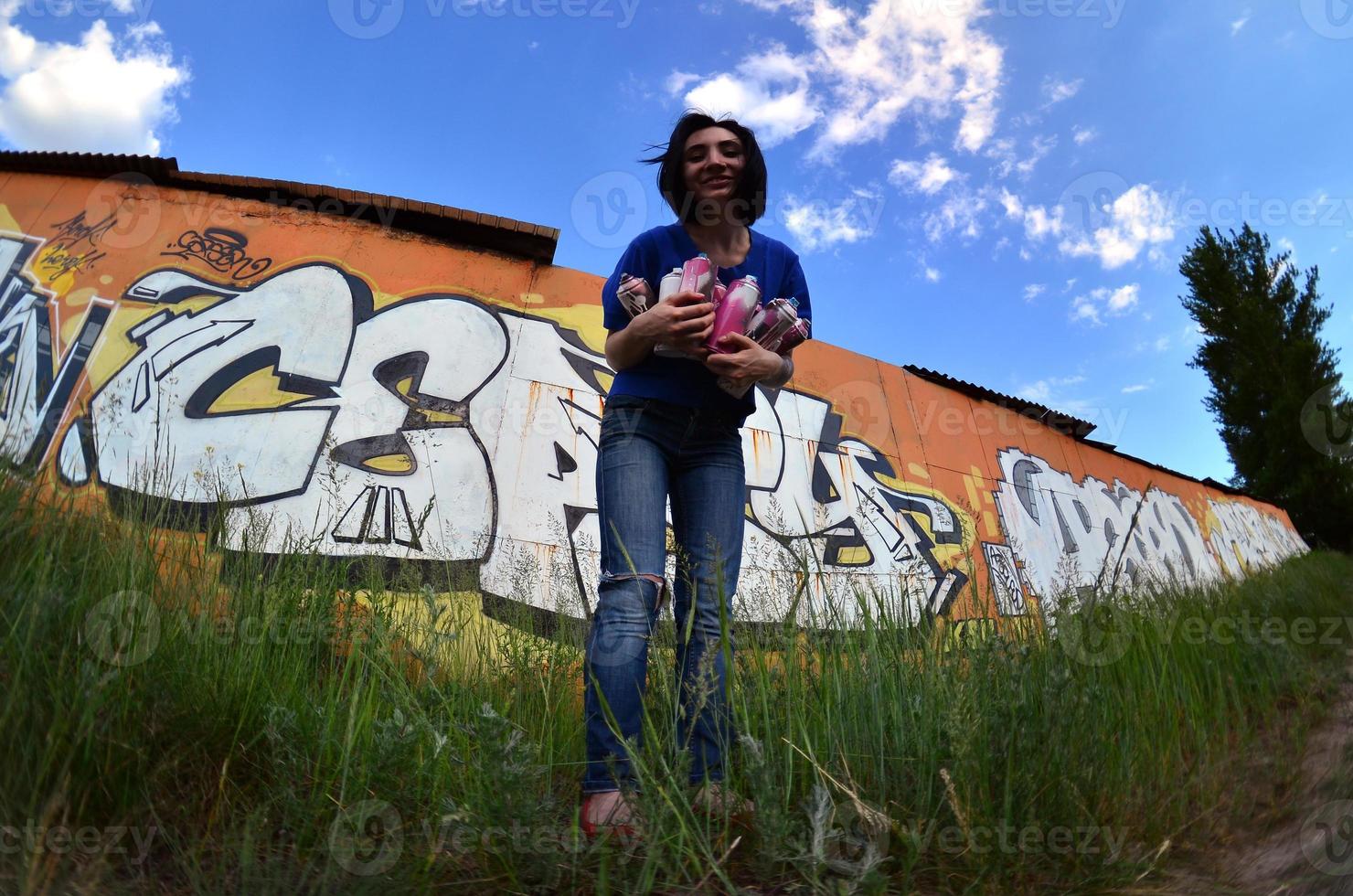 Portrait of an emotional young girl with black hair and piercings. A wide-angle photo of a girl with aerosol paint cans in the hands on a graffiti wall background. A modern portrait of a fisheye lens