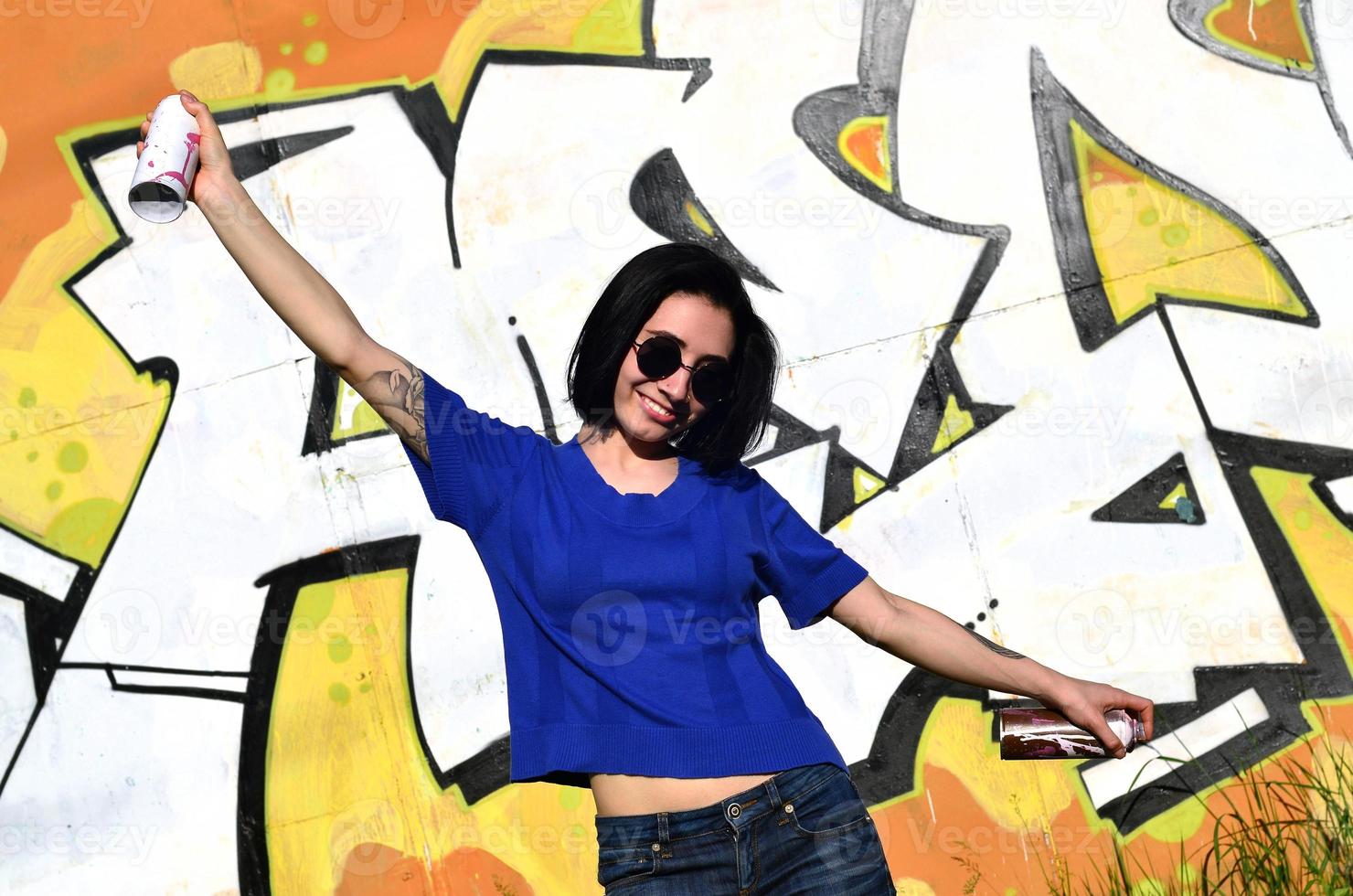 Portrait of an emotional young girl with black hair and piercings. Photo of a girl with aerosol paint cans in hands on a graffiti wall background. The concept of street art and use of aerosol paints