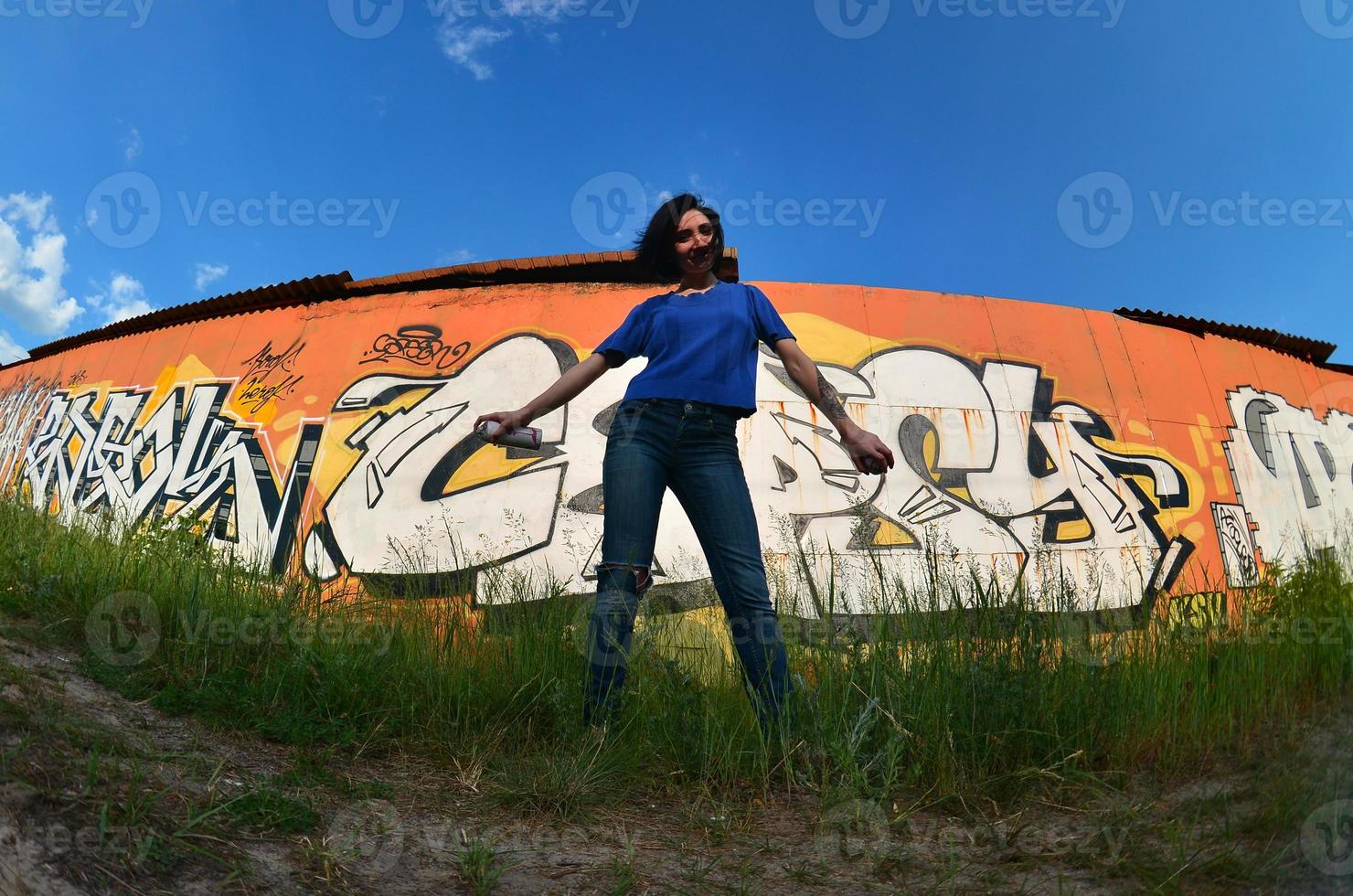 Portrait of an emotional young girl with black hair and piercings. A wide-angle photo of a girl with aerosol paint cans in the hands on a graffiti wall background. A modern portrait of a fisheye lens