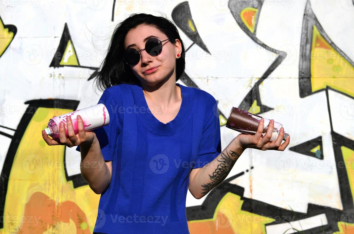 retrato de una joven emocional con pelo negro y piercings. foto de una chica con latas de pintura en aerosol en las manos sobre un fondo de pared de graffiti. el concepto de arte callejero y uso de pinturas en aerosol