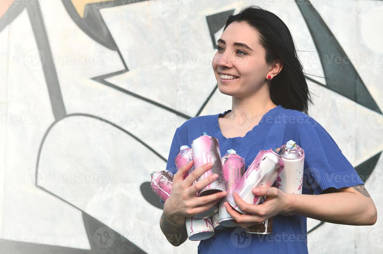 Portrait of an emotional young girl with black hair and piercings. Photo of a girl with aerosol paint cans in hands on a graffiti wall background. The concept of street art and use of aerosol paints