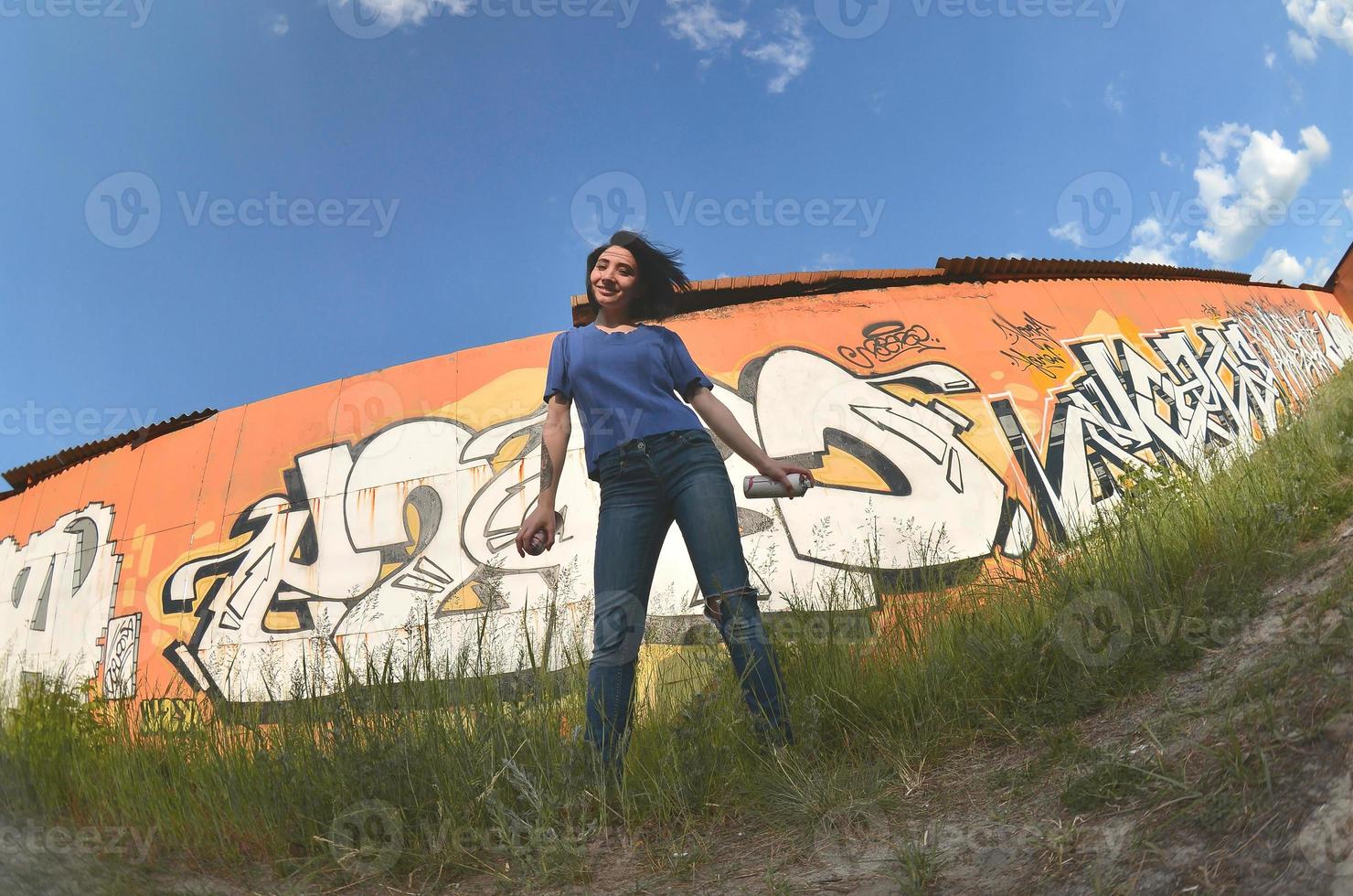 Portrait of an emotional young girl with black hair and piercings. A wide-angle photo of a girl with aerosol paint cans in the hands on a graffiti wall background. A modern portrait of a fisheye lens