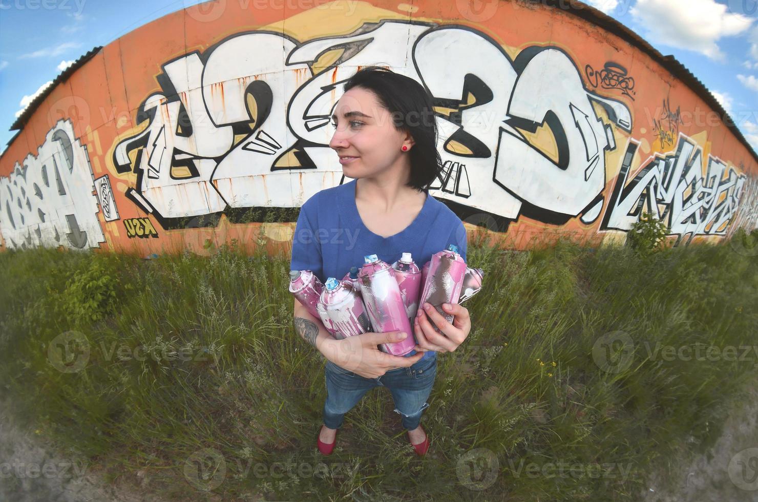 Portrait of an emotional young girl with black hair and piercings. A wide-angle photo of a girl with aerosol paint cans in the hands on a graffiti wall background. A modern portrait of a fisheye lens