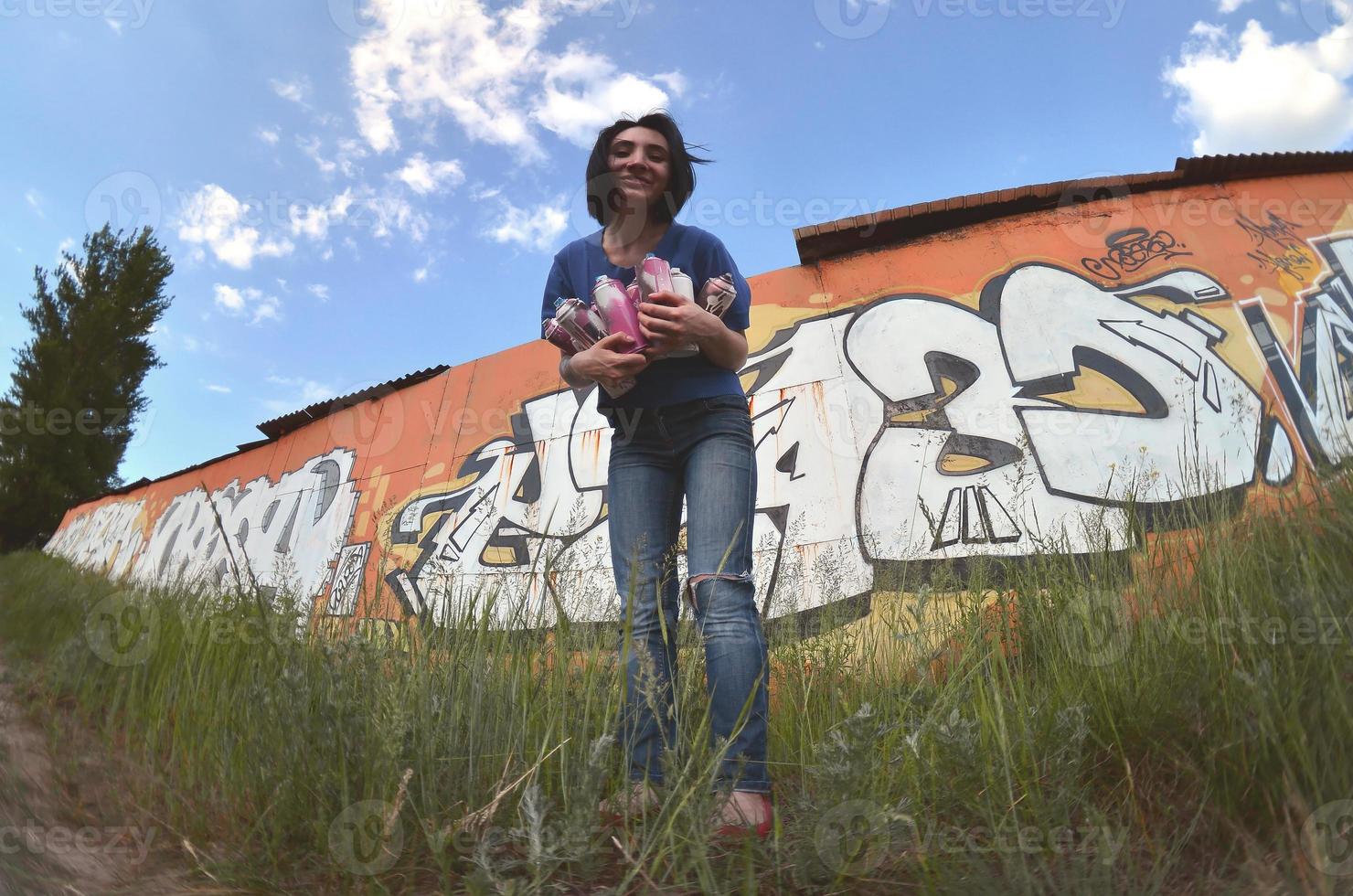 Portrait of an emotional young girl with black hair and piercings. A wide-angle photo of a girl with aerosol paint cans in the hands on a graffiti wall background. A modern portrait of a fisheye lens