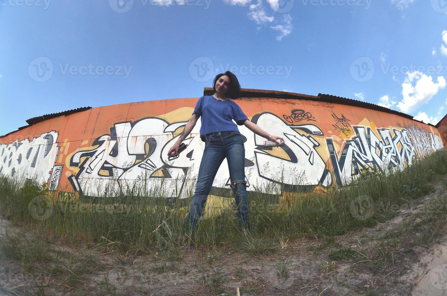 Portrait of an emotional young girl with black hair and piercings. A wide-angle photo of a girl with aerosol paint cans in the hands on a graffiti wall background. A modern portrait of a fisheye lens