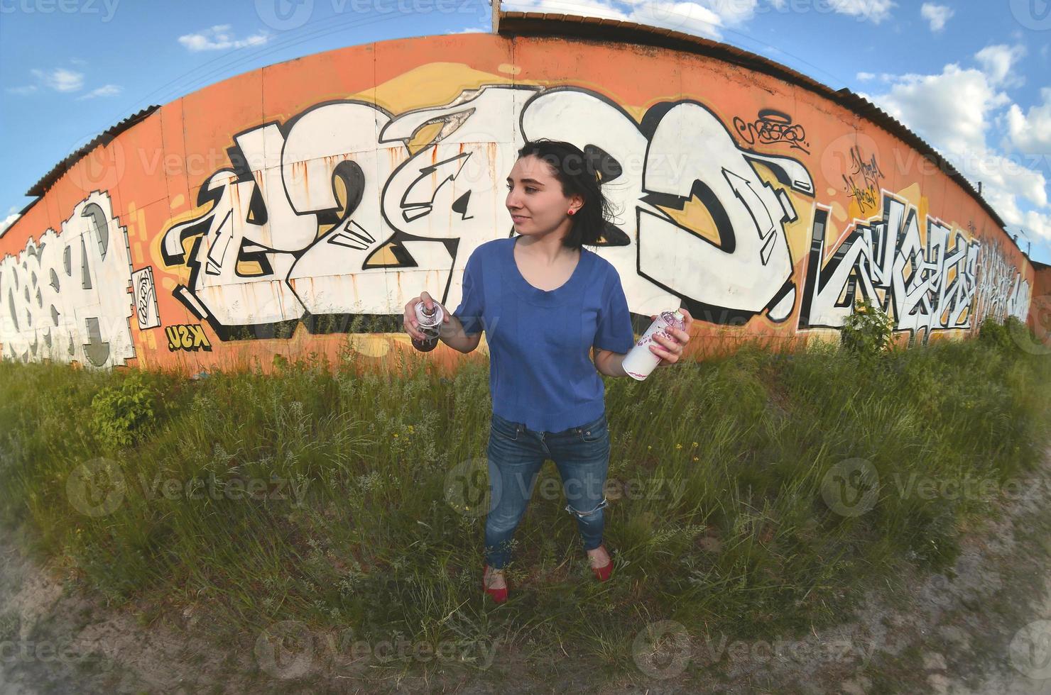 Portrait of an emotional young girl with black hair and piercings. A wide-angle photo of a girl with aerosol paint cans in the hands on a graffiti wall background. A modern portrait of a fisheye lens