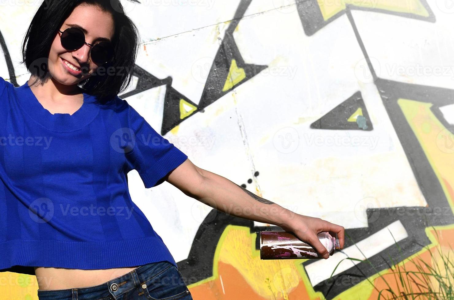 Portrait of an emotional young girl with black hair and piercings. Photo of a girl with aerosol paint cans in hands on a graffiti wall background. The concept of street art and use of aerosol paints