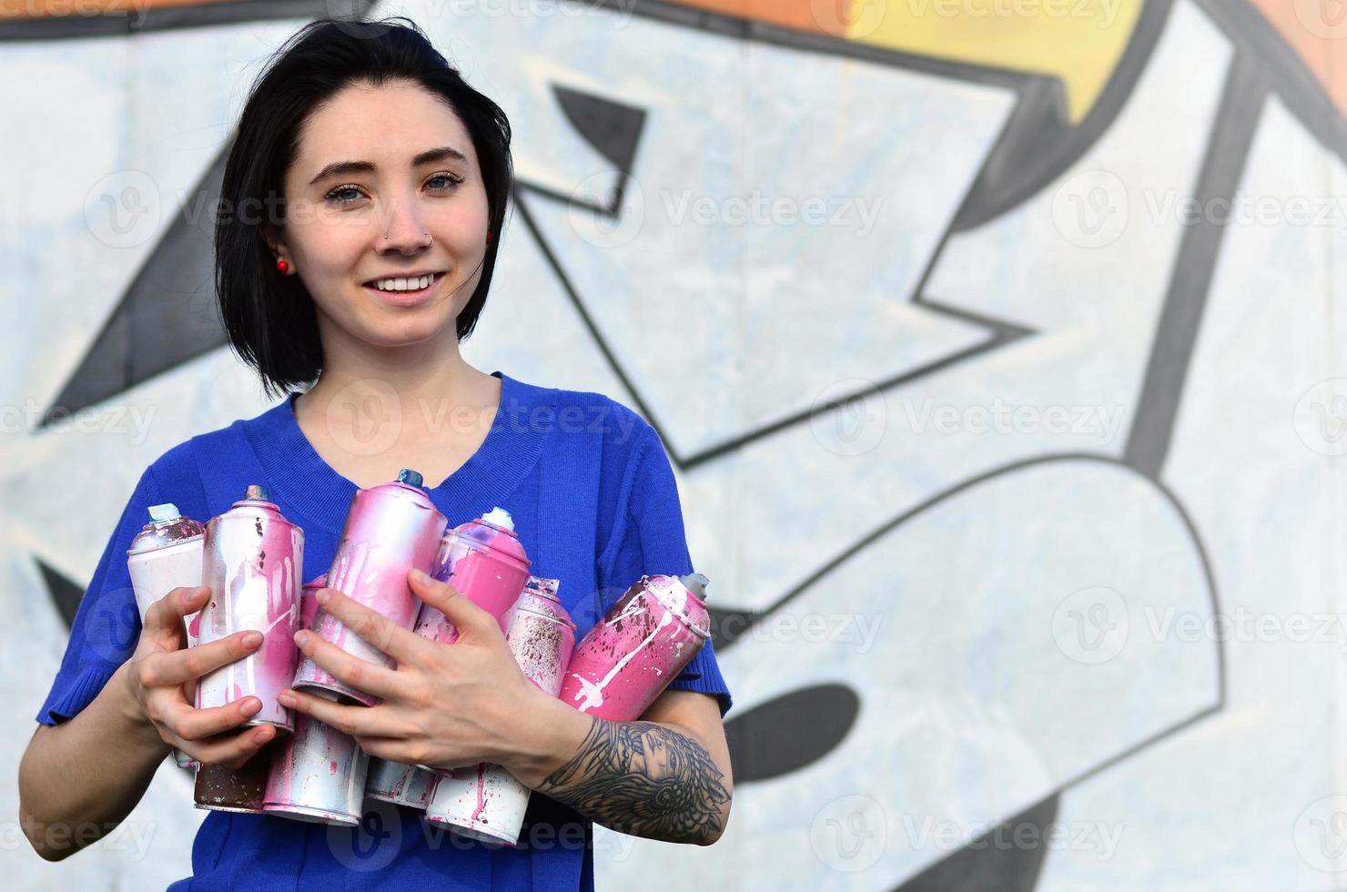 Portrait of an emotional young girl with black hair and piercings. Photo of a girl with aerosol paint cans in hands on a graffiti wall background. The concept of street art and use of aerosol paints
