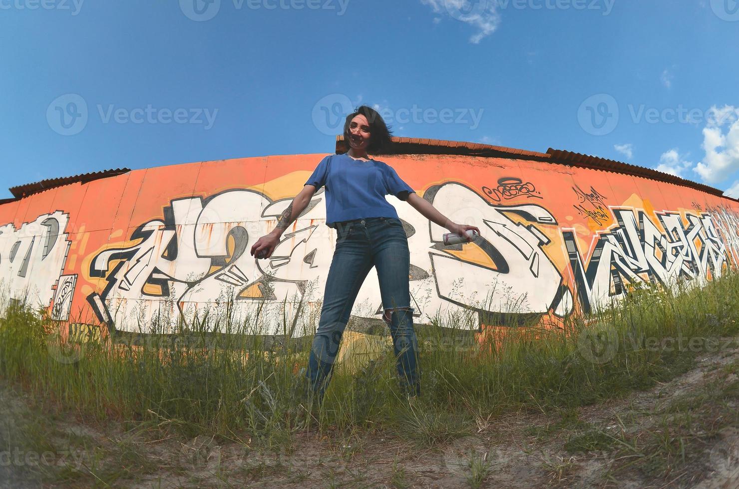 Portrait of an emotional young girl with black hair and piercings. A wide-angle photo of a girl with aerosol paint cans in the hands on a graffiti wall background. A modern portrait of a fisheye lens