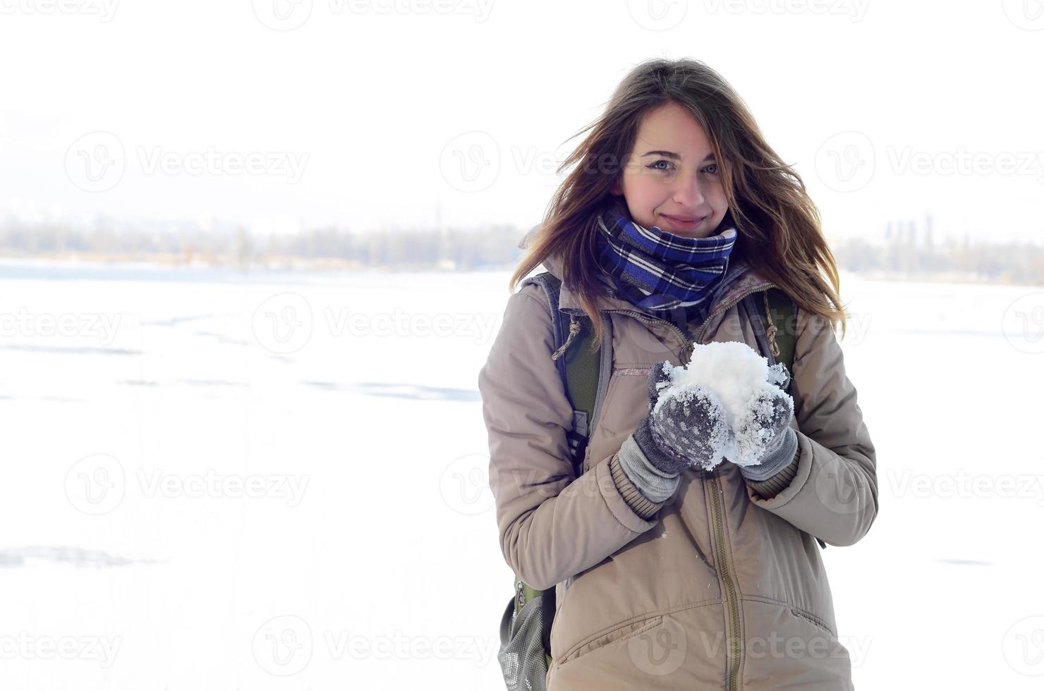 A young and joyful Caucasian girl in a brown coat holds a snowball in the background of a horizon line between the sky and a frozen lake in winter photo