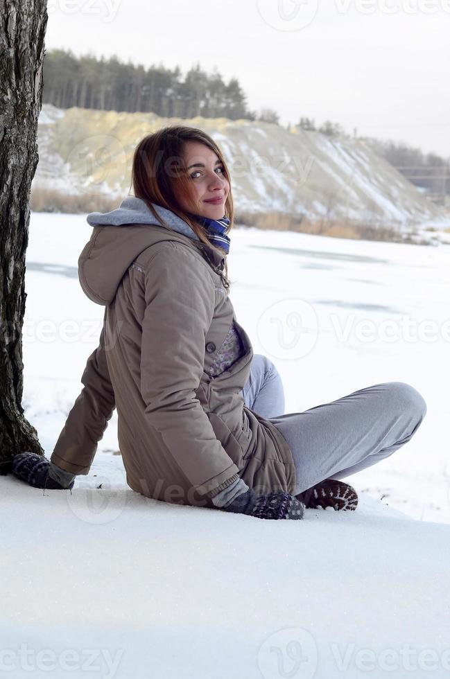 A young Caucasian girl in a brown coat is sitting near a cliff in the background of a horizon line between the sky and a frozen lake in winter time photo