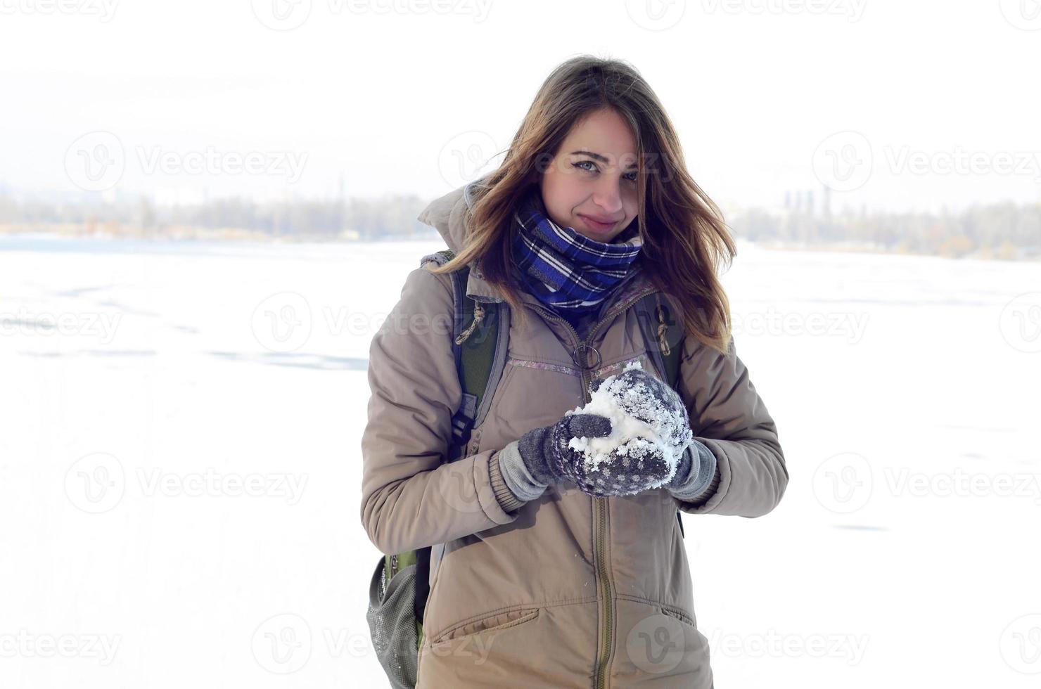 A young and joyful Caucasian girl in a brown coat holds a snowball in the background of a horizon line between the sky and a frozen lake in winter photo