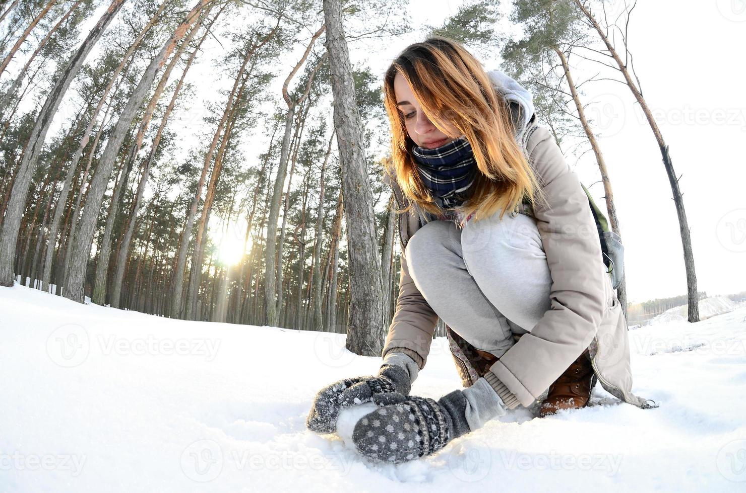 una joven y alegre chica caucásica con un abrigo marrón esculpe una bola de nieve en un bosque cubierto de nieve en invierno. juegos con nieve al aire libre. foto de ojo de pez