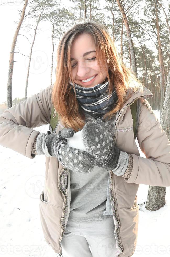 una joven y alegre chica caucásica con un abrigo marrón esculpe una bola de nieve en un bosque cubierto de nieve en invierno. juegos con nieve al aire libre. foto de ojo de pez