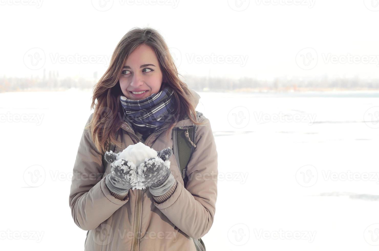 A young and joyful Caucasian girl in a brown coat holds a snowball in the background of a horizon line between the sky and a frozen lake in winter photo