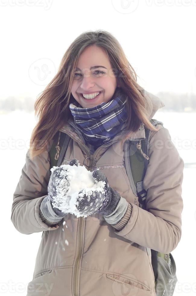 A young and joyful Caucasian girl in a brown coat holds a snowball in the background of a horizon line between the sky and a frozen lake in winter photo