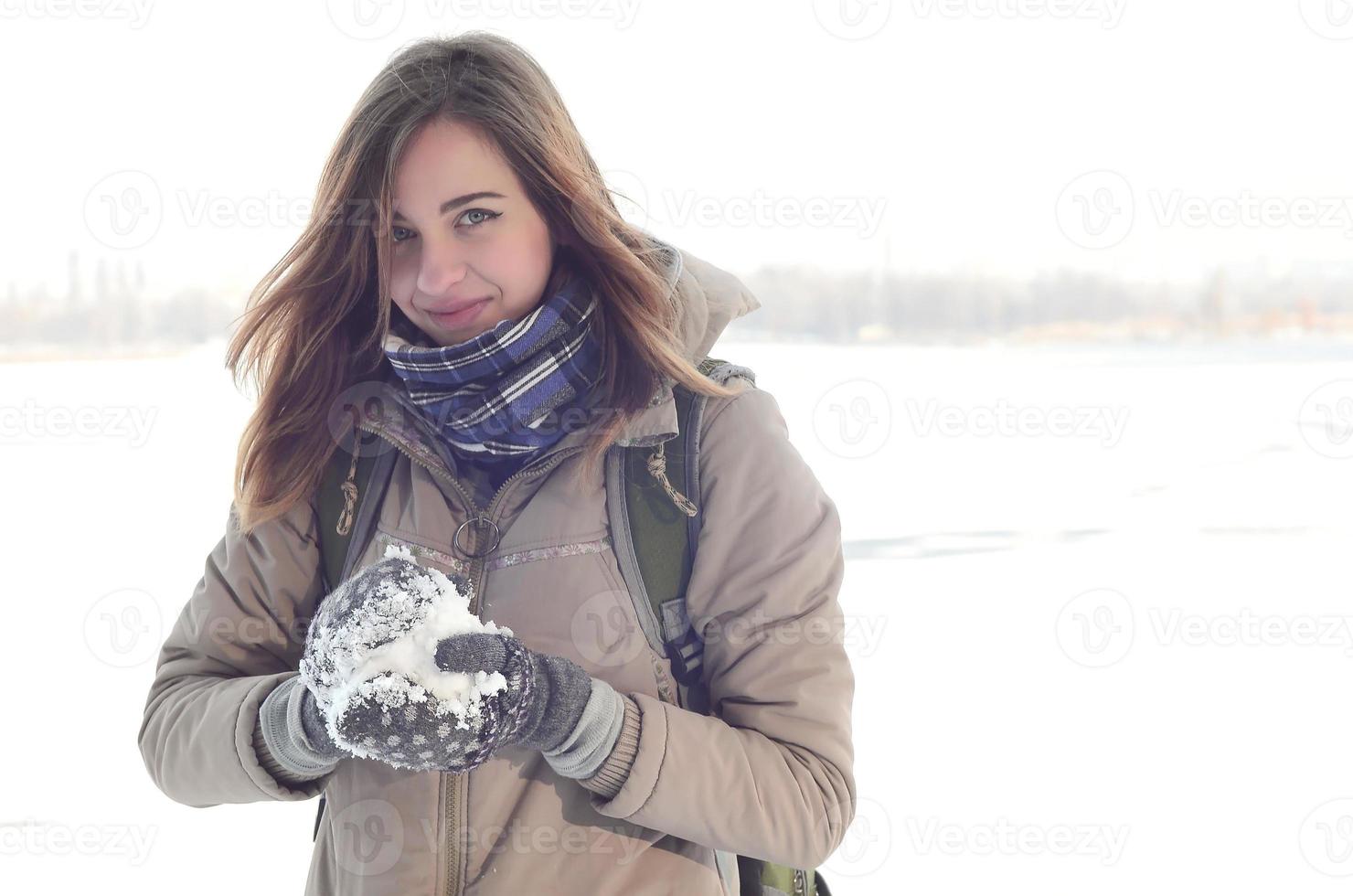 A young and joyful Caucasian girl in a brown coat holds a snowball in the background of a horizon line between the sky and a frozen lake in winter photo