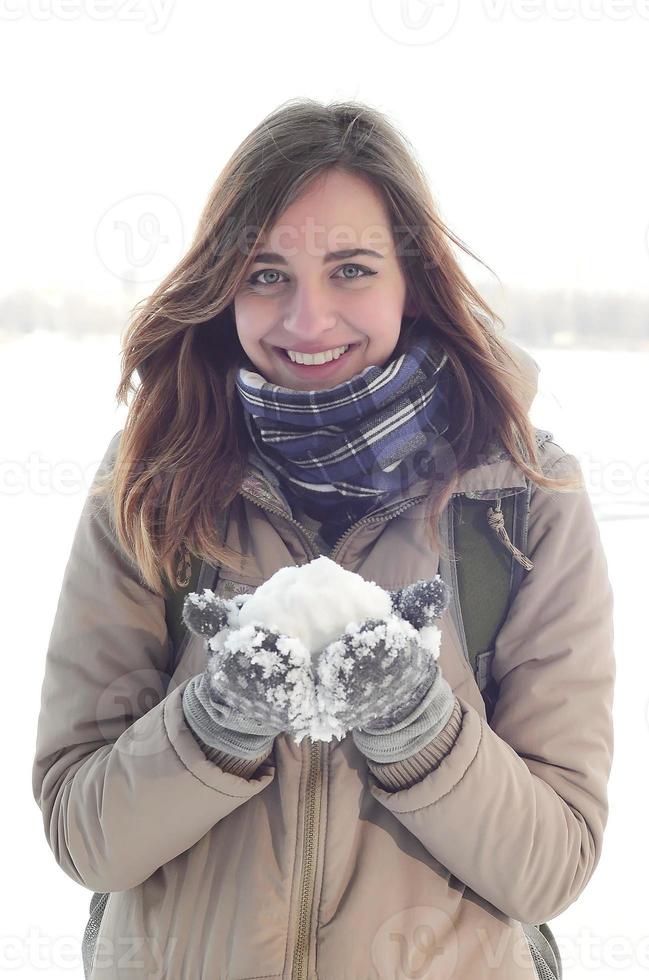 A young and joyful Caucasian girl in a brown coat holds a snowball in the background of a horizon line between the sky and a frozen lake in winter photo