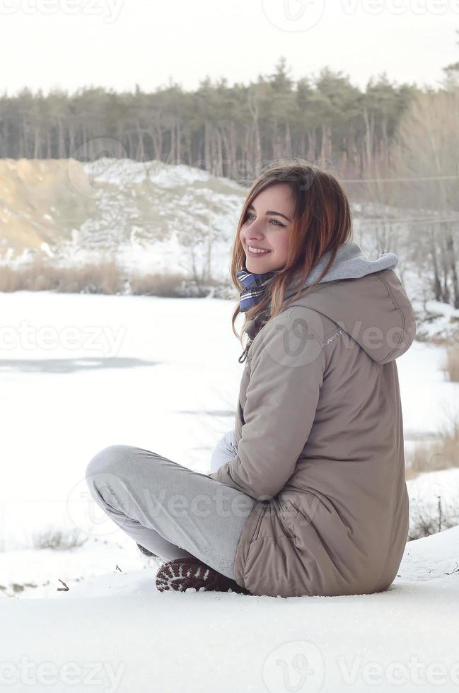 A young Caucasian girl in a brown coat is sitting near a cliff in the background of a horizon line between the sky and a frozen lake in winter time photo