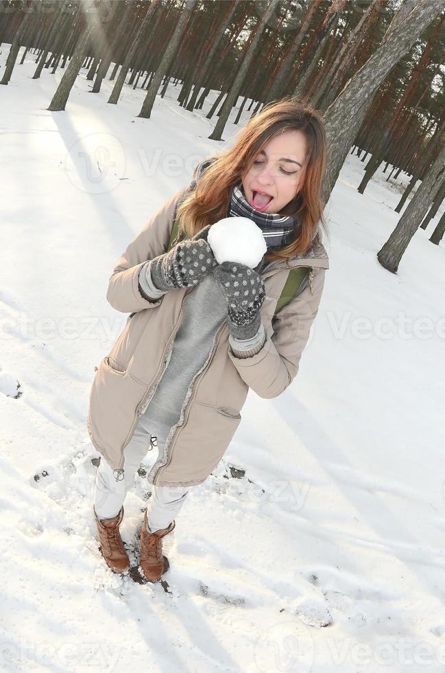 A young and joyful Caucasian girl in a brown coat holds a snowball in a snow-covered forest in winter. Fisheye Photo