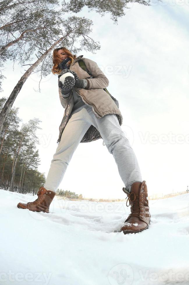 A young and joyful Caucasian girl in a brown coat holds a snowball in front of a horizon line between the sky and a frozen lake in winter. Fisheye Photo