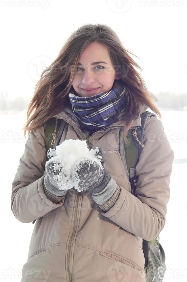 A young and joyful Caucasian girl in a brown coat holds a snowball in the background of a horizon line between the sky and a frozen lake in winter photo