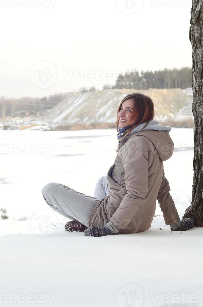 A young Caucasian girl in a brown coat is sitting near a cliff in the background of a horizon line between the sky and a frozen lake in winter time photo