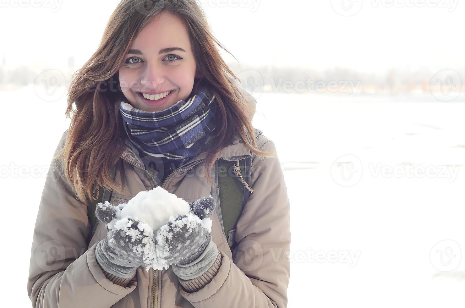 A young and joyful Caucasian girl in a brown coat holds a snowball in the background of a horizon line between the sky and a frozen lake in winter photo