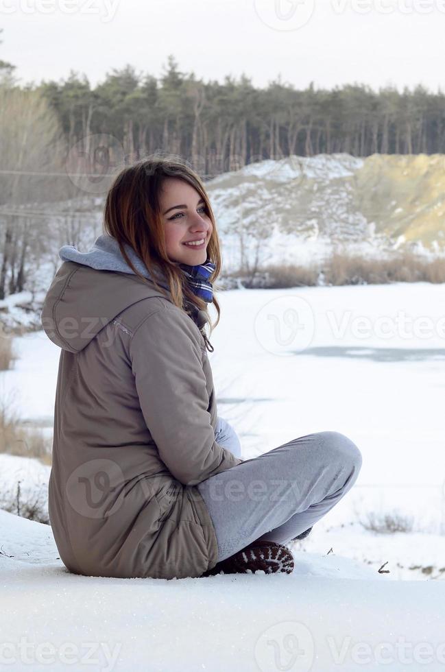 A young Caucasian girl in a brown coat is sitting near a cliff in the background of a horizon line between the sky and a frozen lake in winter time photo