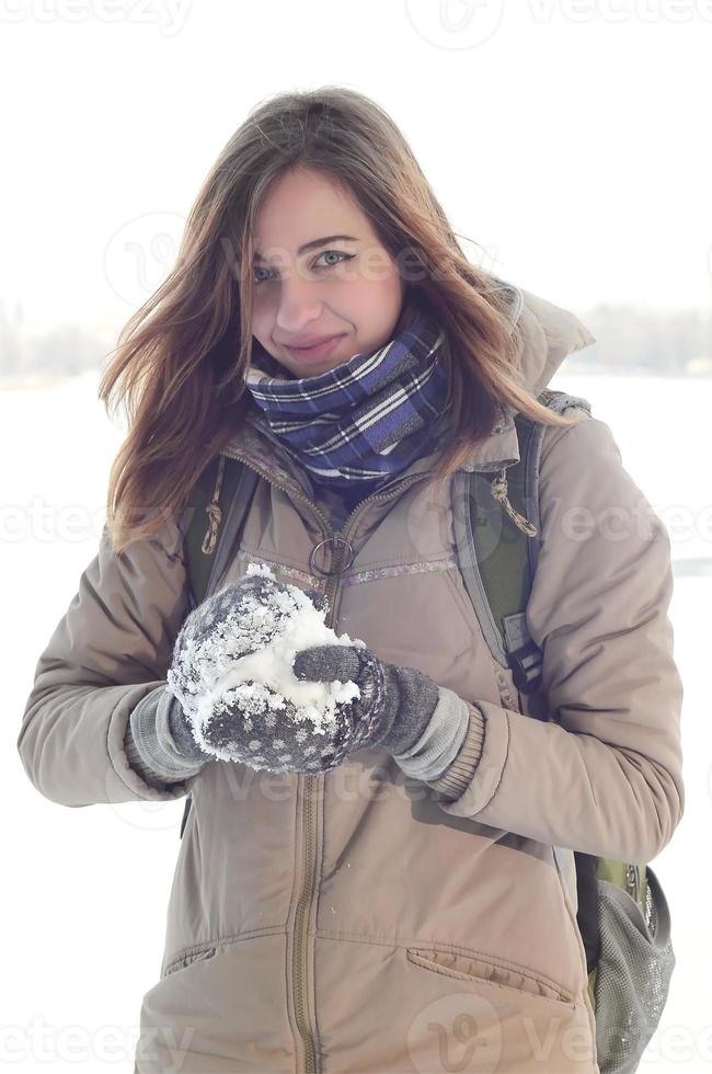 A young and joyful Caucasian girl in a brown coat holds a snowball in the background of a horizon line between the sky and a frozen lake in winter photo