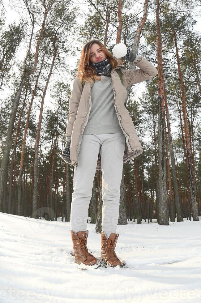 una joven y alegre chica caucásica con un abrigo marrón sostiene una bola de nieve en un bosque cubierto de nieve en invierno. foto de ojo de pez