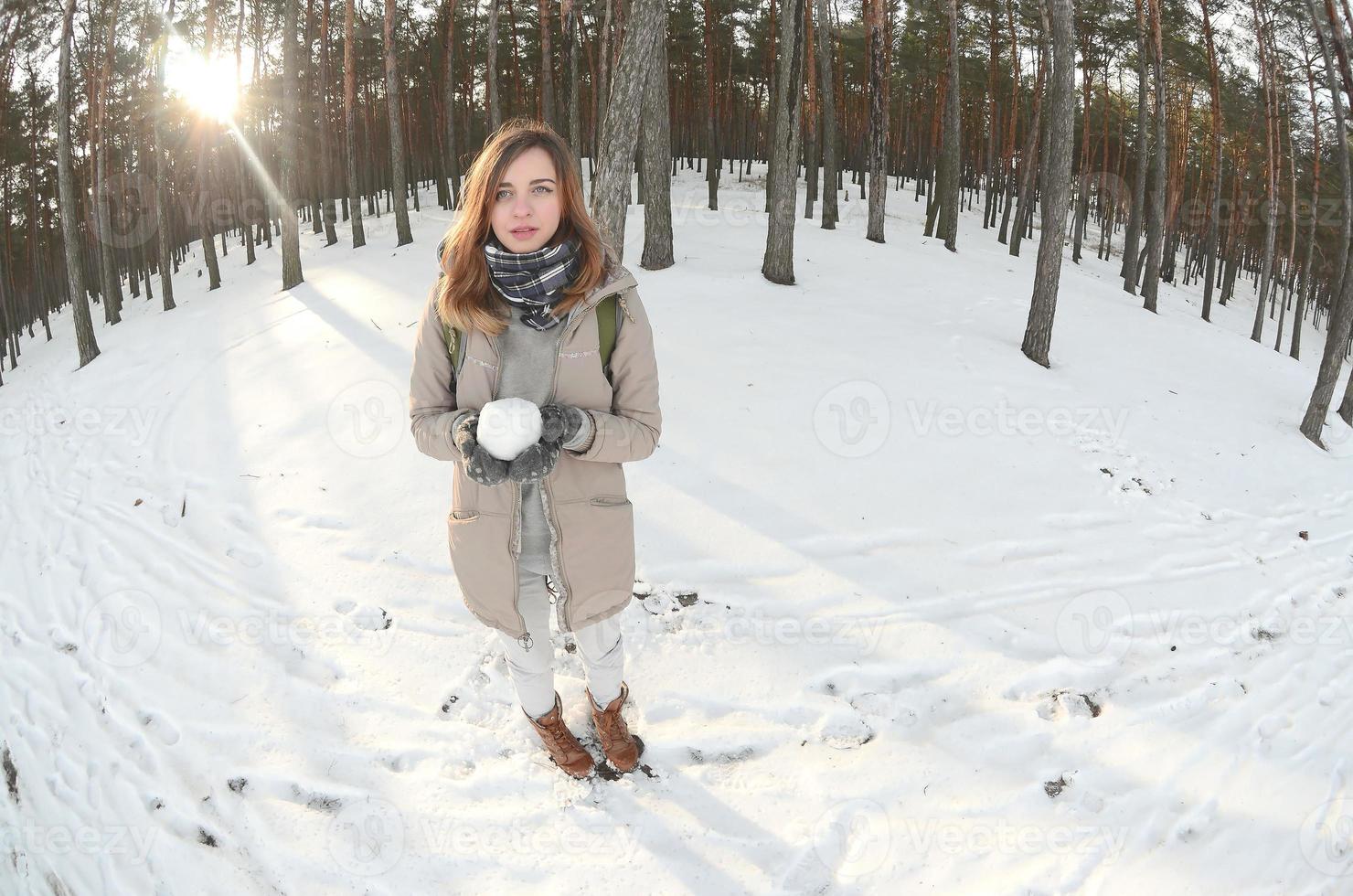 A young and joyful Caucasian girl in a brown coat holds a snowball in a snow-covered forest in winter. Fisheye Photo