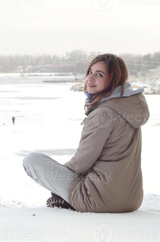 A young Caucasian girl in a brown coat is sitting near a cliff in the background of a horizon line between the sky and a frozen lake in winter time photo