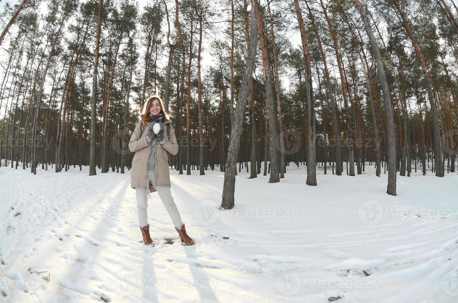 A young and joyful Caucasian girl in a brown coat holds a snowball in a snow-covered forest in winter. Fisheye Photo