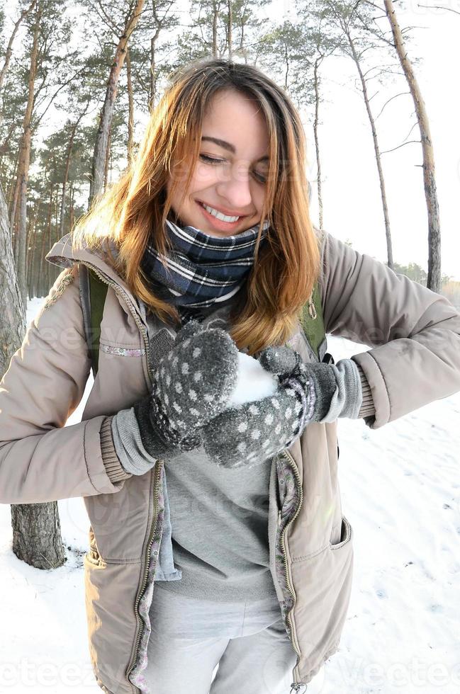 A young and joyful Caucasian girl in a brown coat sculpts a snowball in a snow-covered forest in winter. Games with snow in the open air. Fisheye Photo