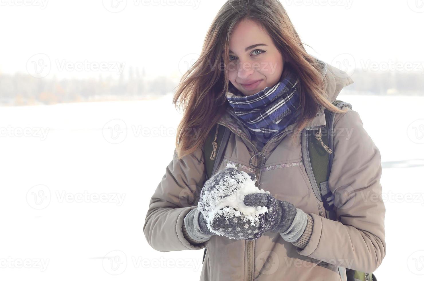 A young and joyful Caucasian girl in a brown coat holds a snowball in the background of a horizon line between the sky and a frozen lake in winter photo