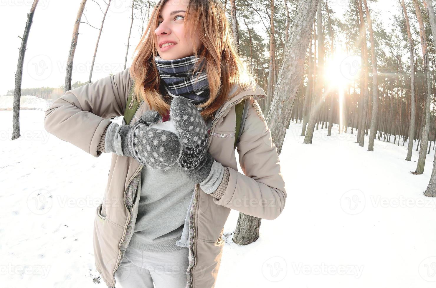 una joven y alegre chica caucásica con un abrigo marrón esculpe una bola de nieve en un bosque cubierto de nieve en invierno. juegos con nieve al aire libre. foto de ojo de pez