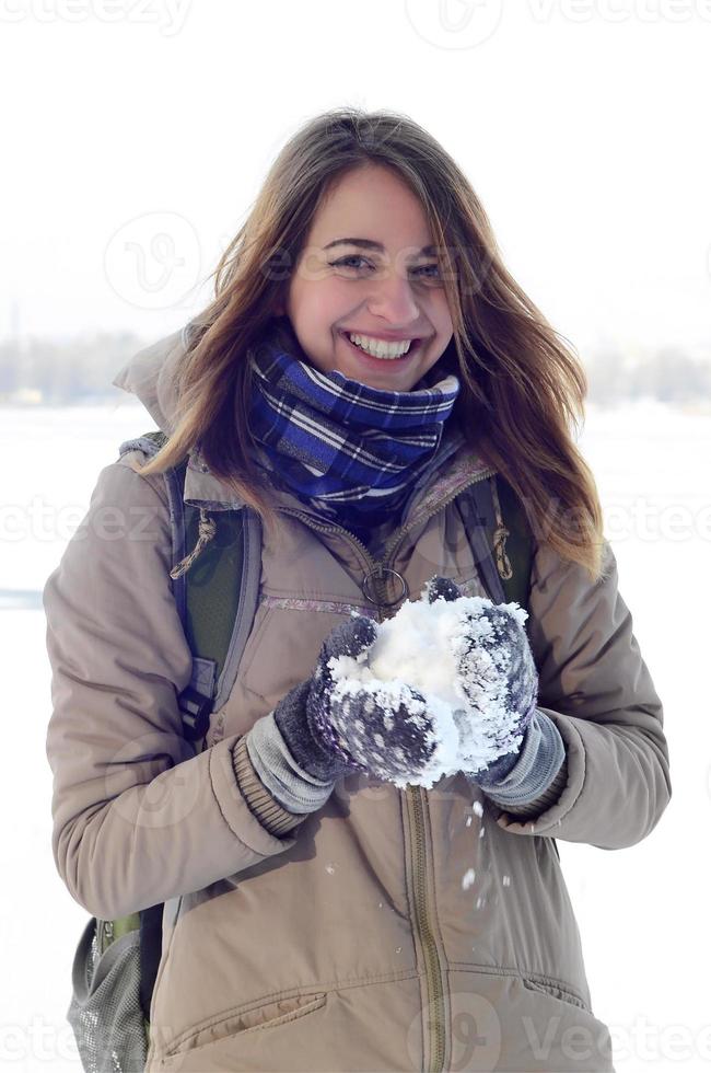 A young and joyful Caucasian girl in a brown coat holds a snowball in the background of a horizon line between the sky and a frozen lake in winter photo