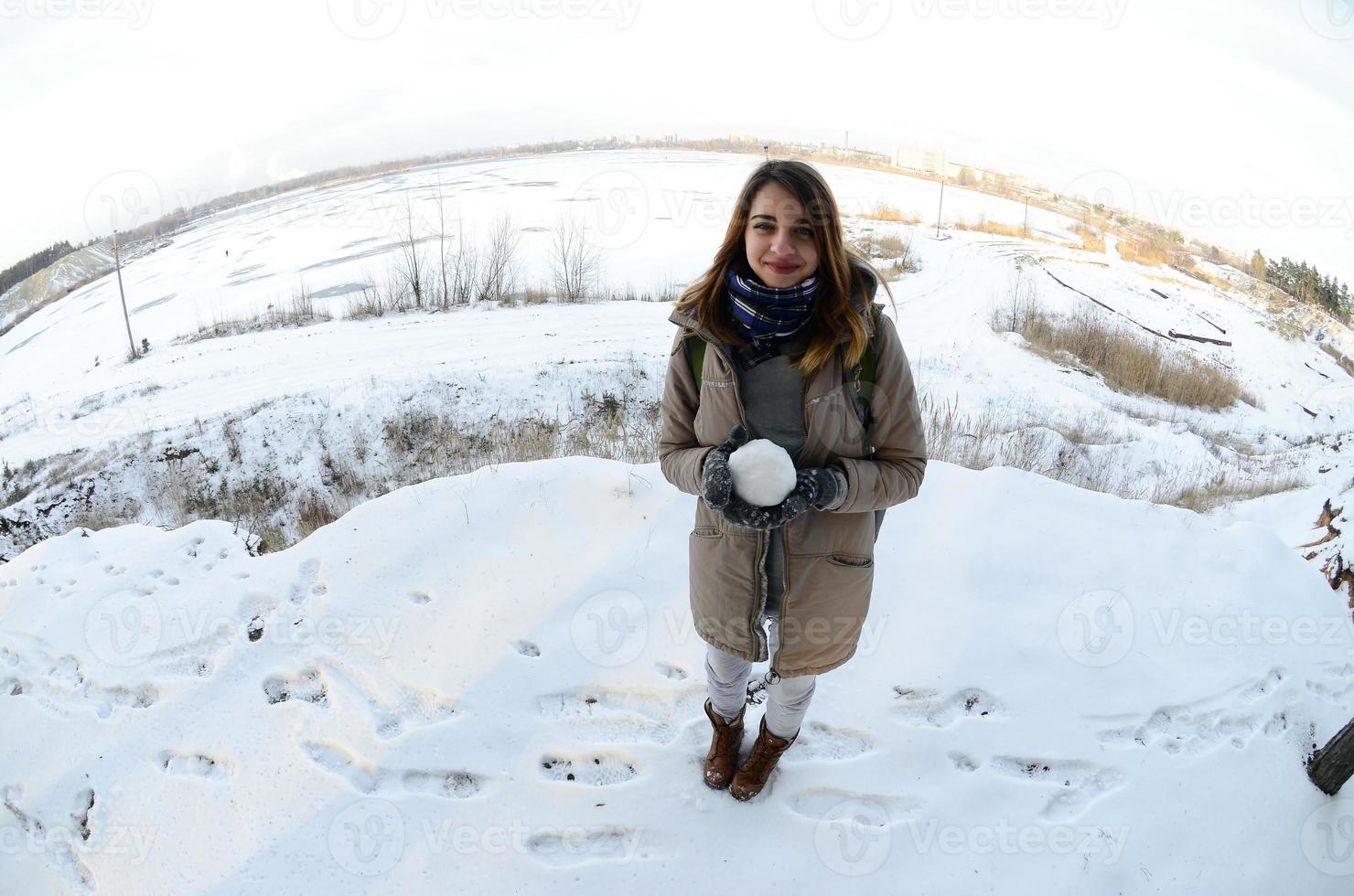 A young and joyful Caucasian girl in a brown coat holds a snowball in front of a horizon line between the sky and a frozen lake in winter. Fisheye Photo