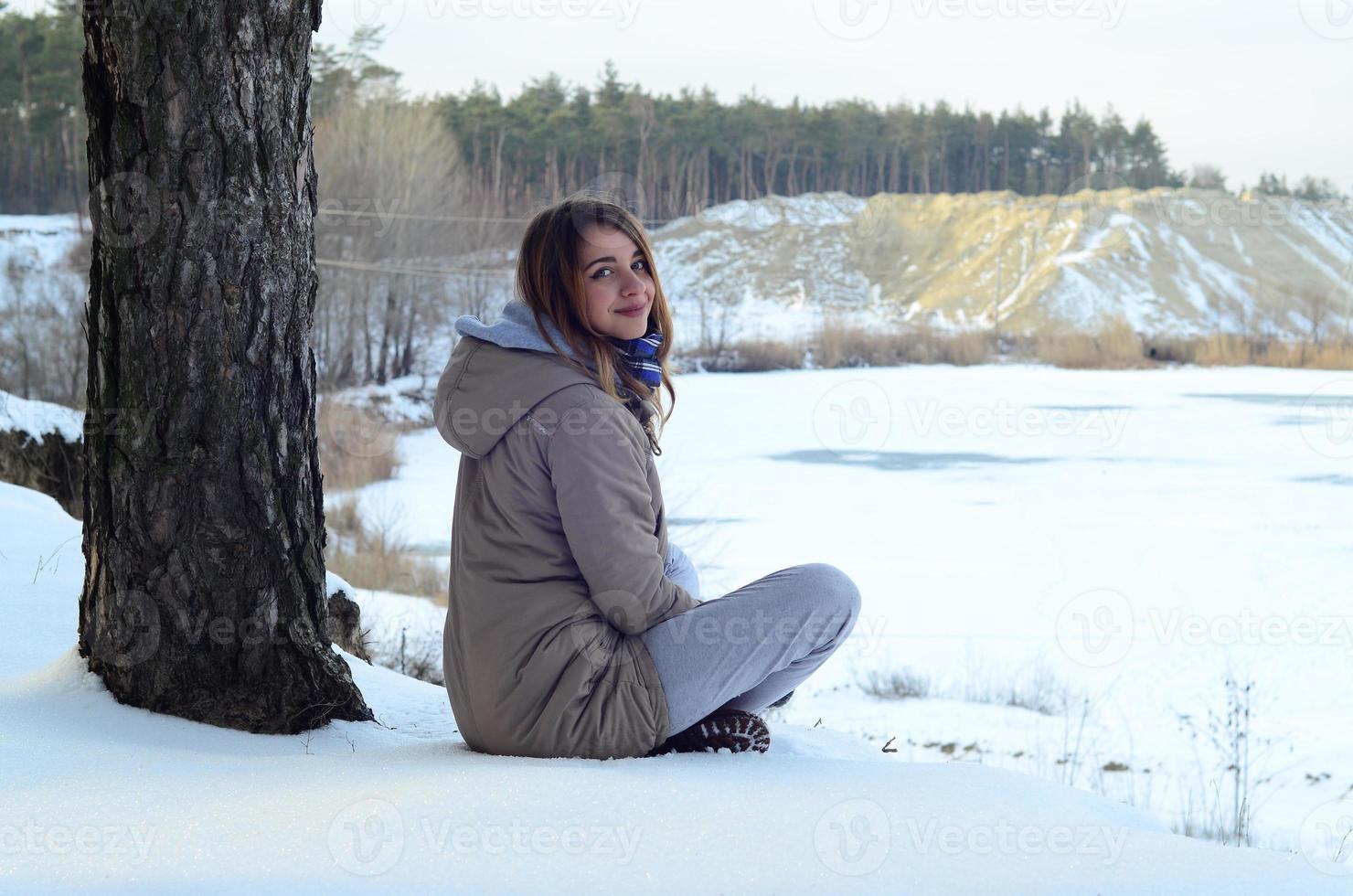 A young Caucasian girl in a brown coat is sitting near a cliff in the background of a horizon line between the sky and a frozen lake in winter time photo