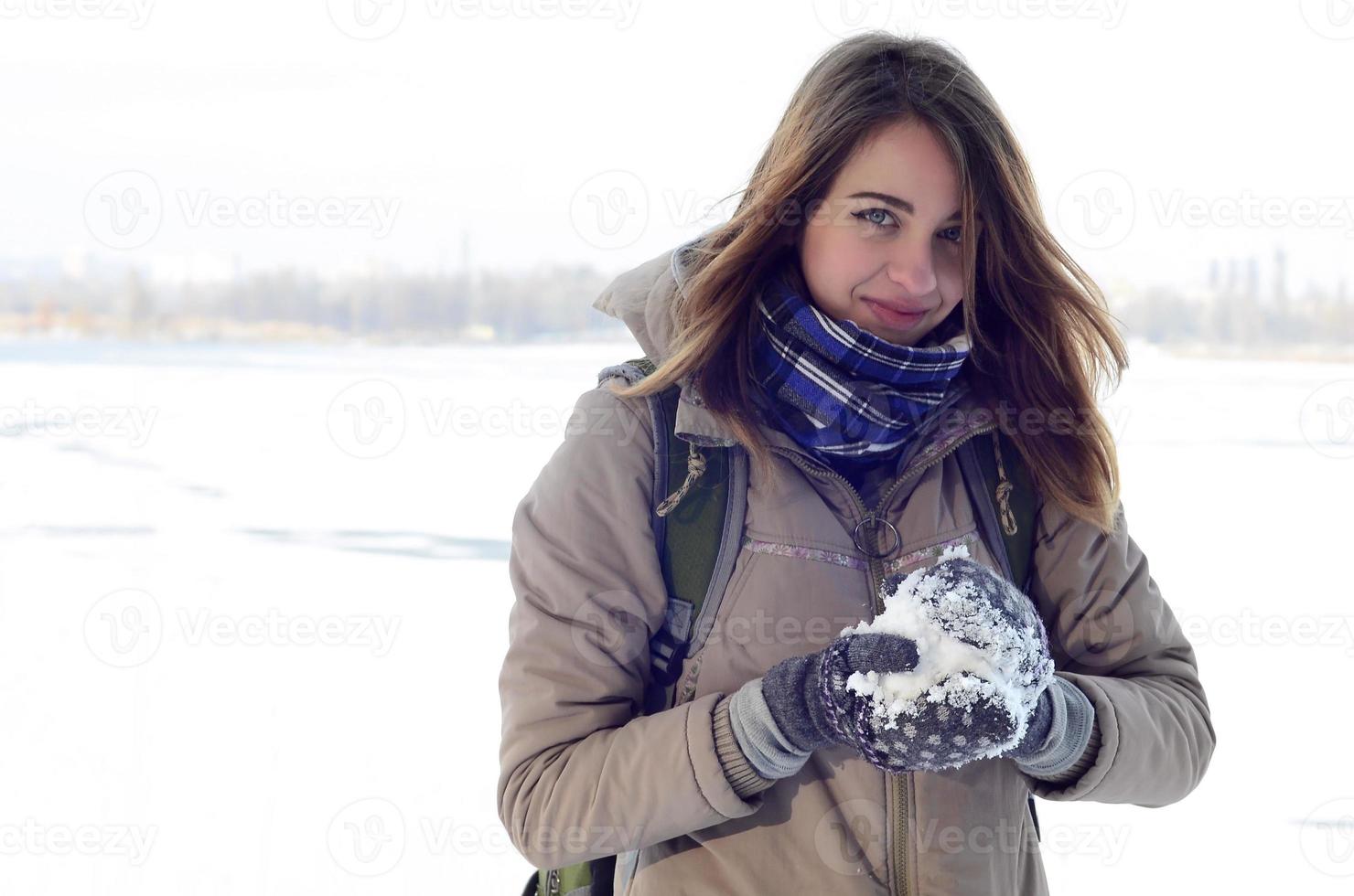 A young and joyful Caucasian girl in a brown coat holds a snowball in the background of a horizon line between the sky and a frozen lake in winter photo