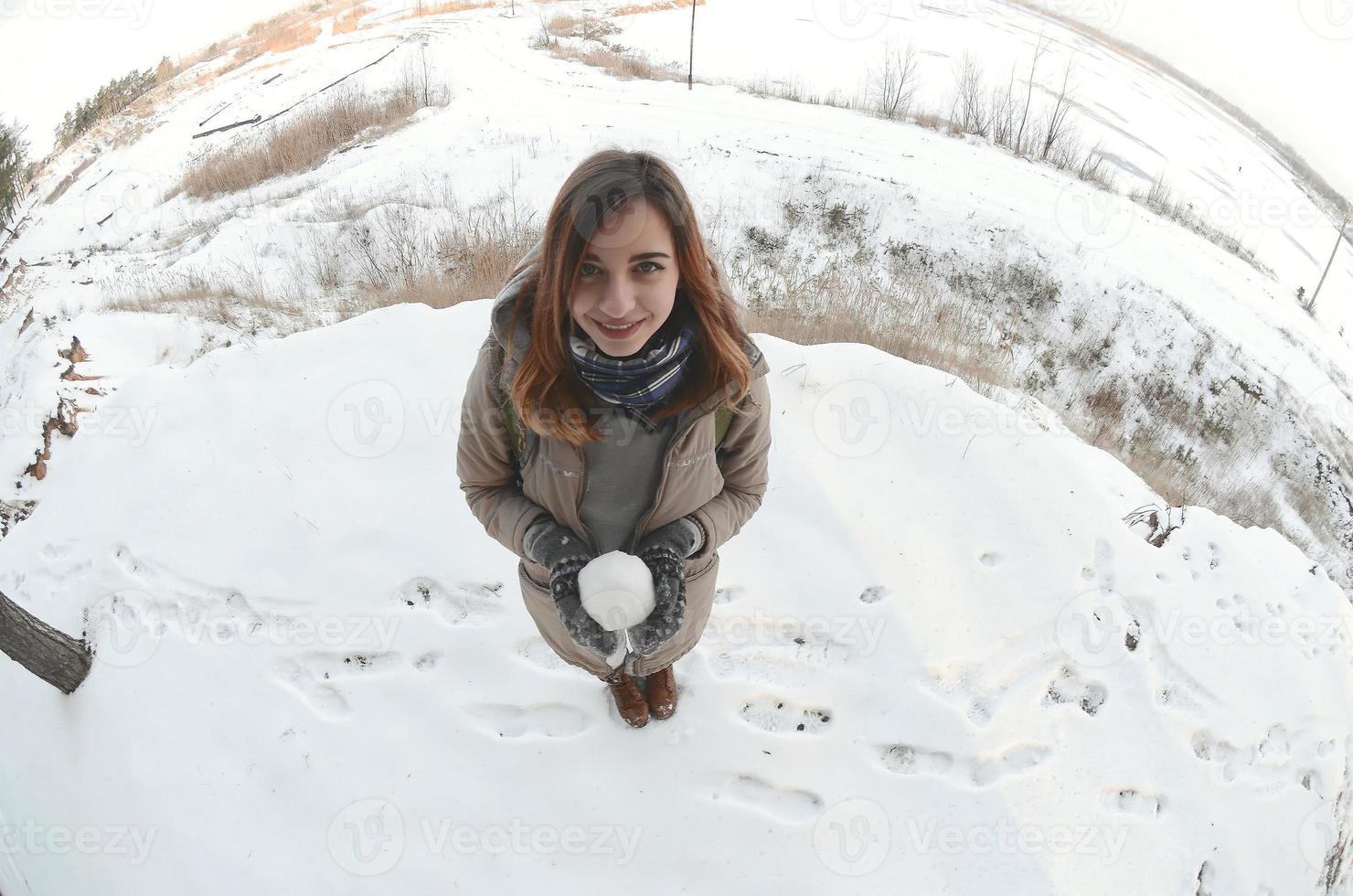 A young and joyful Caucasian girl in a brown coat holds a snowball in front of a horizon line between the sky and a frozen lake in winter. Fisheye Photo