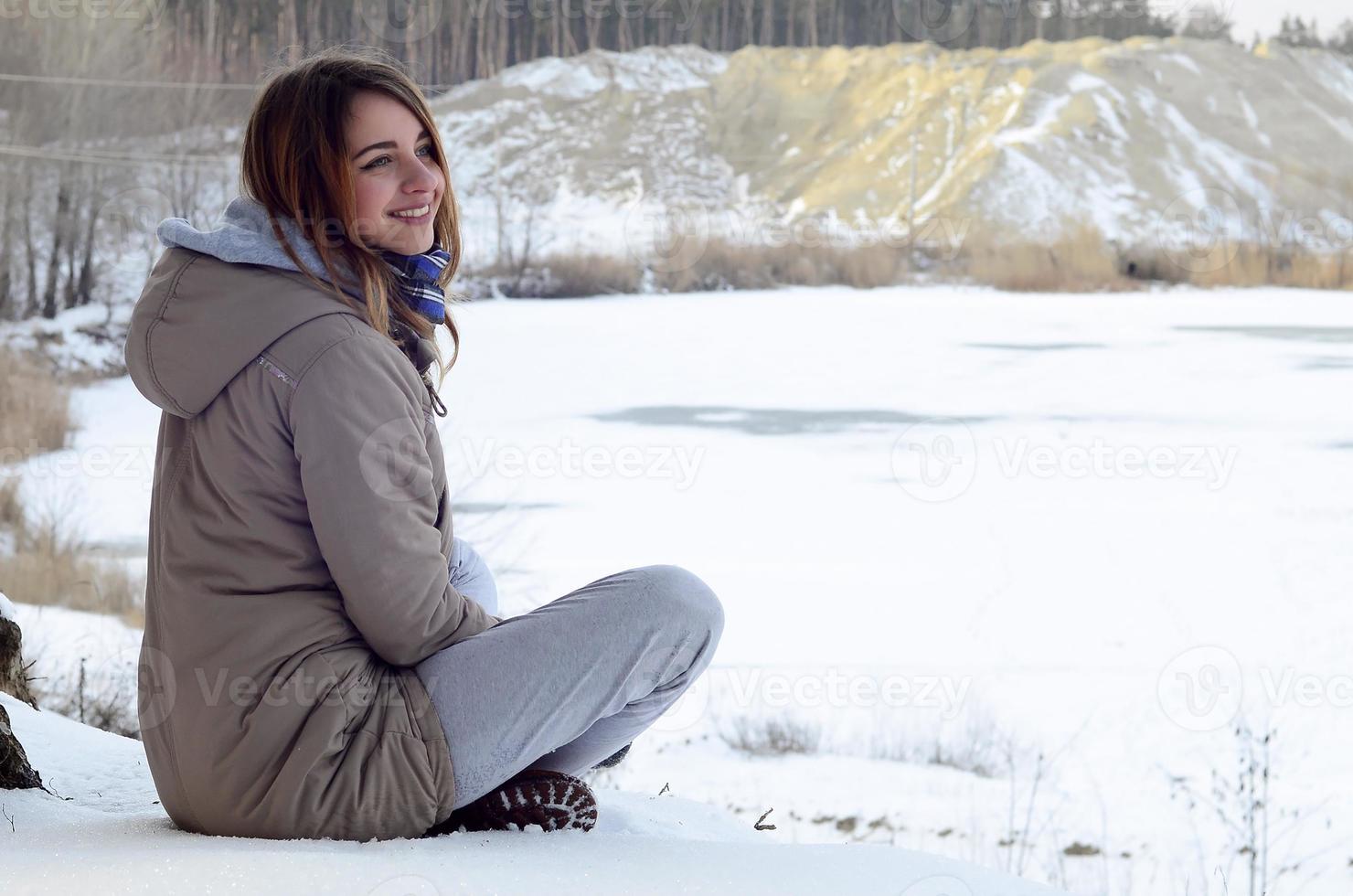 A young Caucasian girl in a brown coat is sitting near a cliff in the background of a horizon line between the sky and a frozen lake in winter time photo