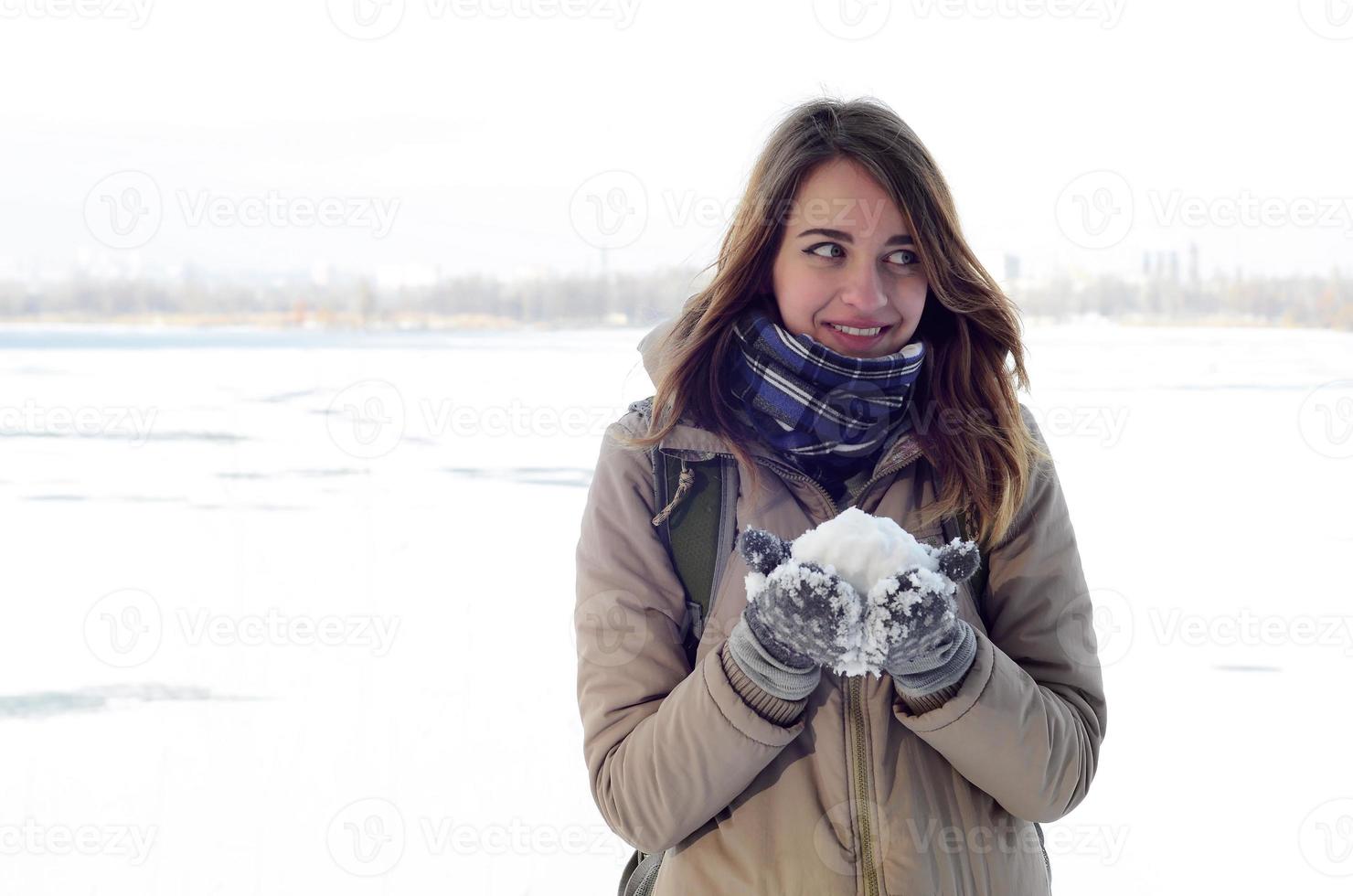 A young and joyful Caucasian girl in a brown coat holds a snowball in the background of a horizon line between the sky and a frozen lake in winter photo
