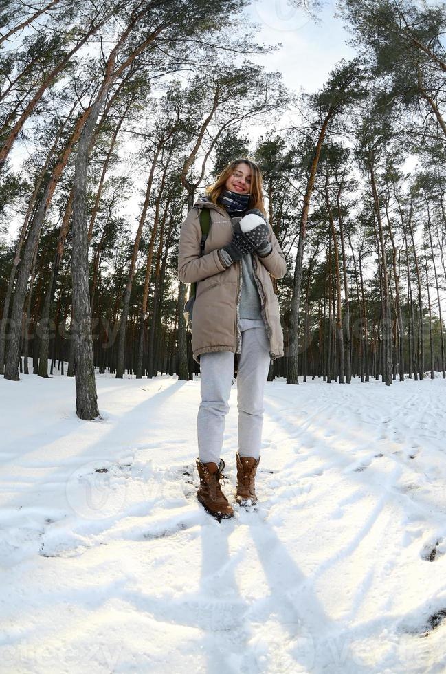 una joven y alegre chica caucásica con un abrigo marrón sostiene una bola de nieve en un bosque cubierto de nieve en invierno. foto de ojo de pez