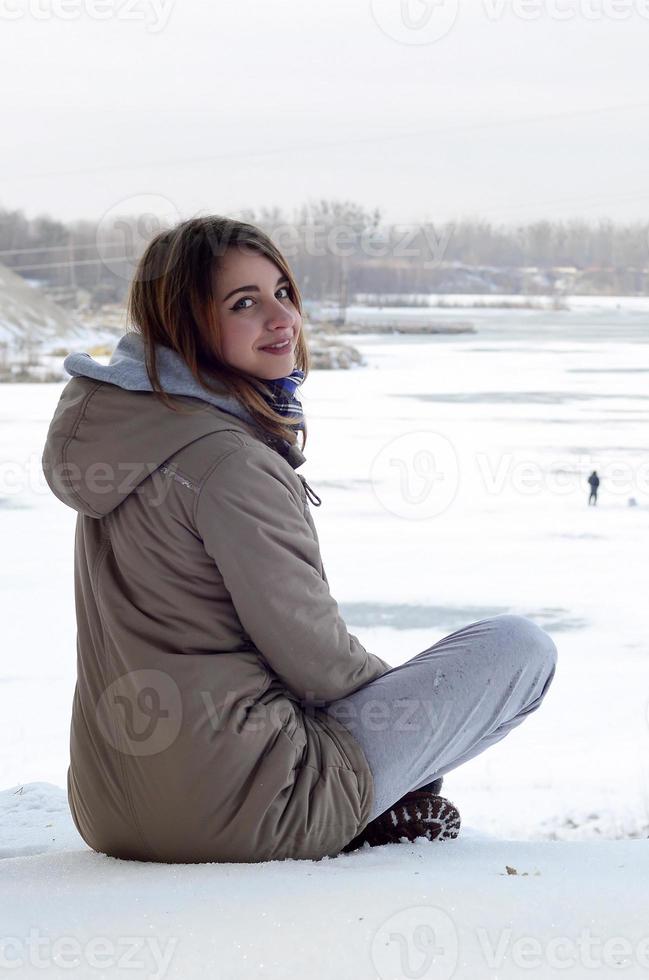 A young Caucasian girl in a brown coat is sitting near a cliff in the background of a horizon line between the sky and a frozen lake in winter time photo